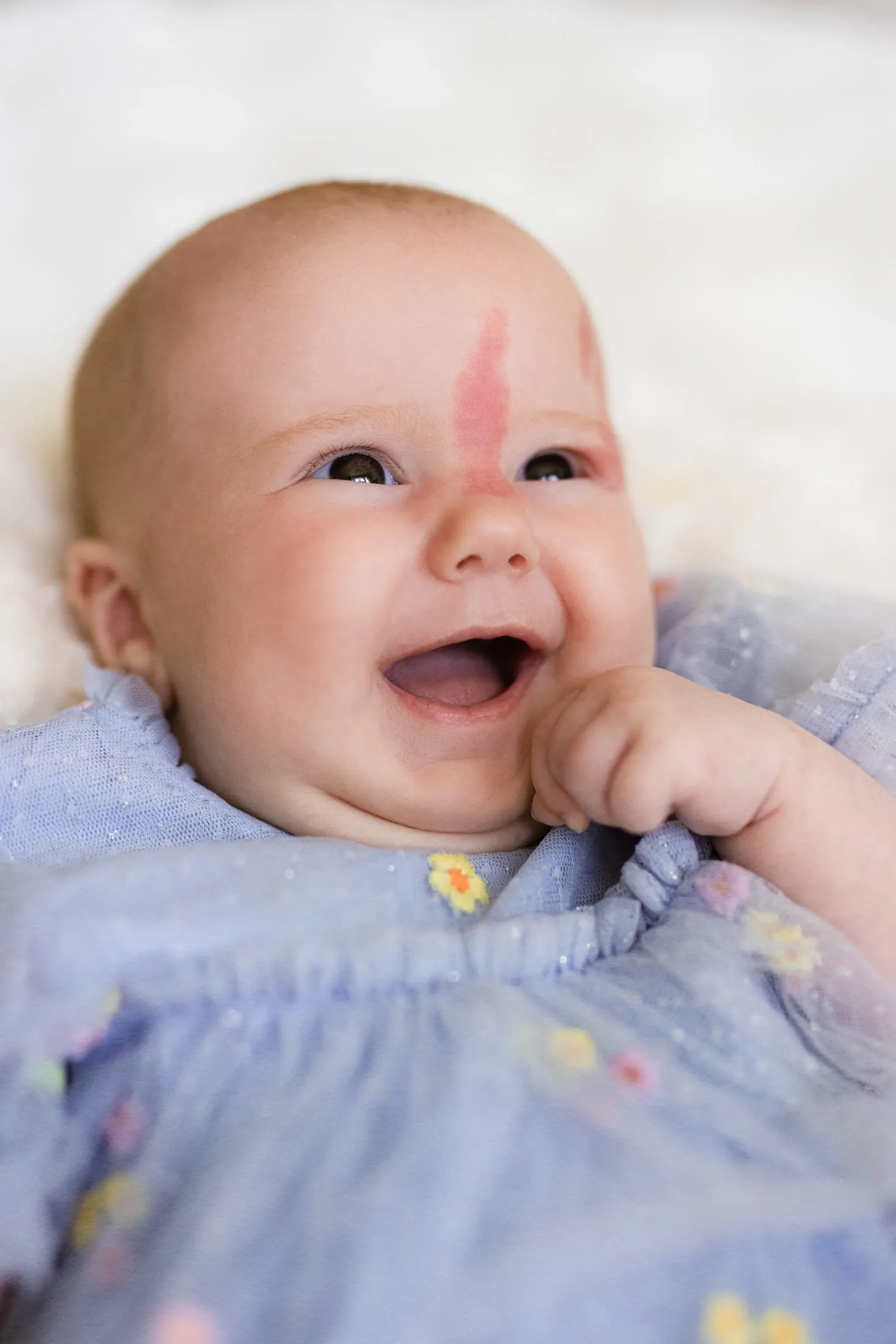 A laughing baby in a blue floral dress on a soft white blanket