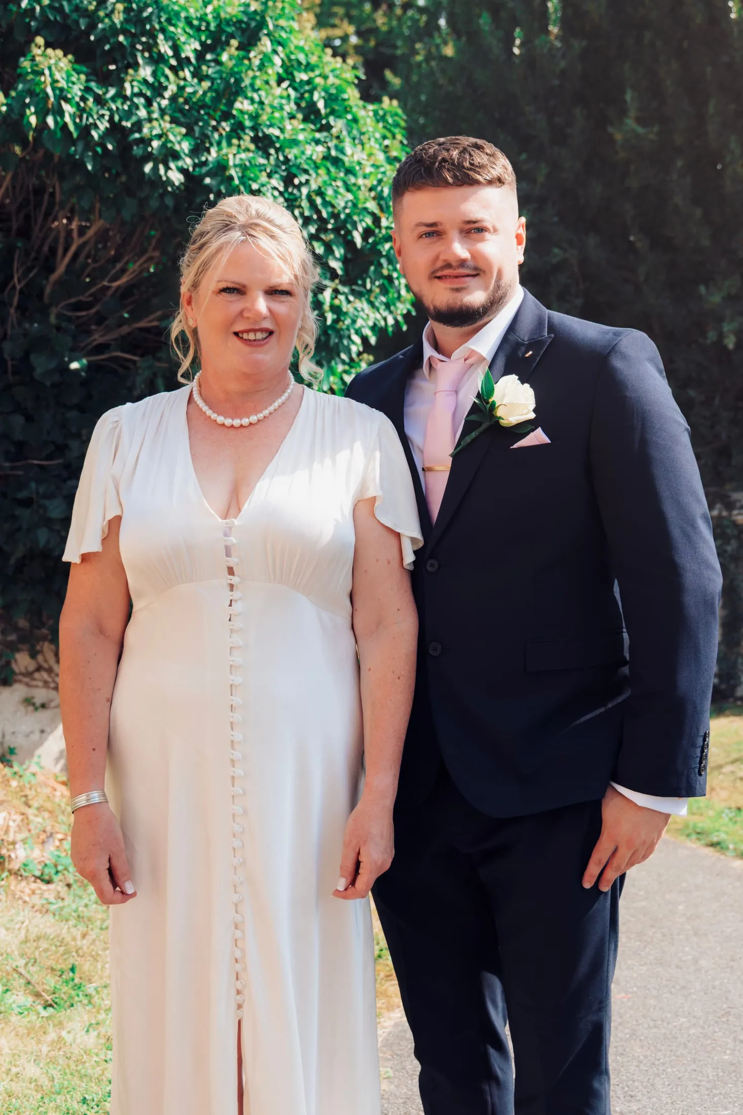 Bride with a young man in a navy suit and rose buttonhole in a sunlit garden